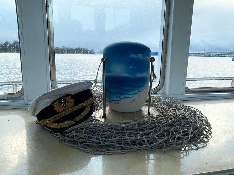 Seebestattungsurne mit nautischer Deko vor einem Schiffsfenster arrangiert.