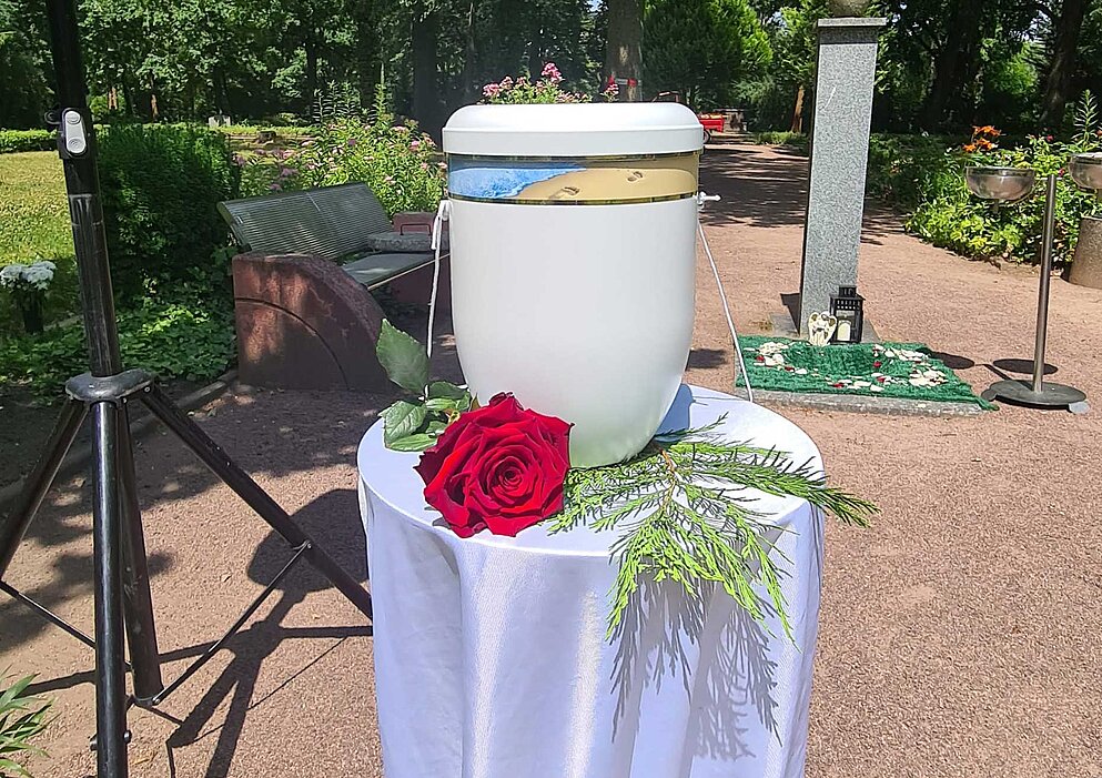 Weiße Urne mit roter Rose dekoriert aufgebahrt auf Tisch mit weißem Tuch.