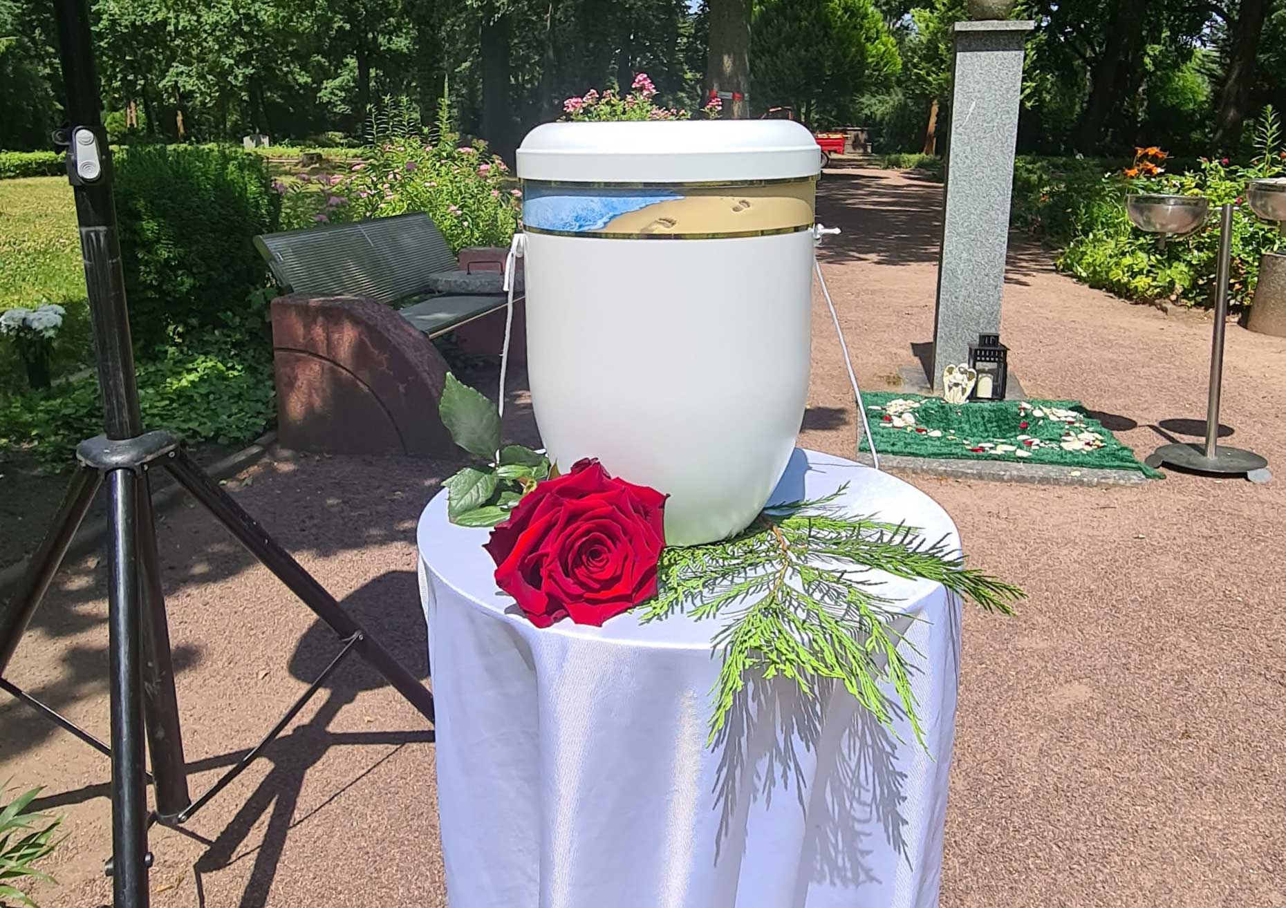 Weiße Urne mit roter Rose dekoriert aufgebahrt auf Tisch mit weißem Tuch.
